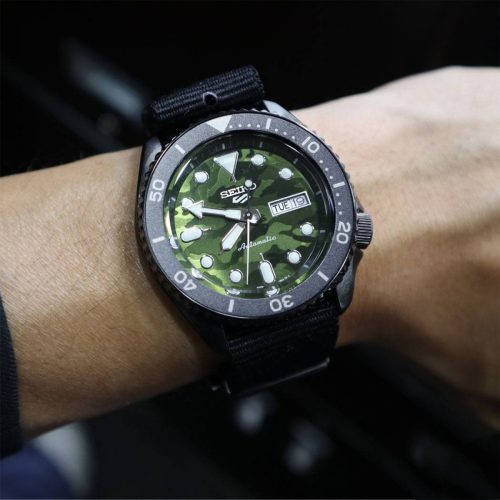 Seiko SRPJ37K1 5 Sports Camouflage SKX sat na ruci muškarca, sa kamuflažnim brojčanikom i crnom najlon narukvicom. Sat je savršen za sportske aktivnosti i outdoor avanture