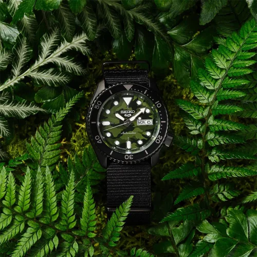 Seiko SRPJ37K1 5 Sports Camouflage SKX muški ručni sat sa kamuflažnim brojčanikom, tamno srebrnim čeličnim kućištem i crnom najlon tekstilnom narukvicom. Sat sa automatskim mehanizmom i dodatnim funkcijama dana u nedelji i datum. Sa slikan u sumi u zelenom liscu