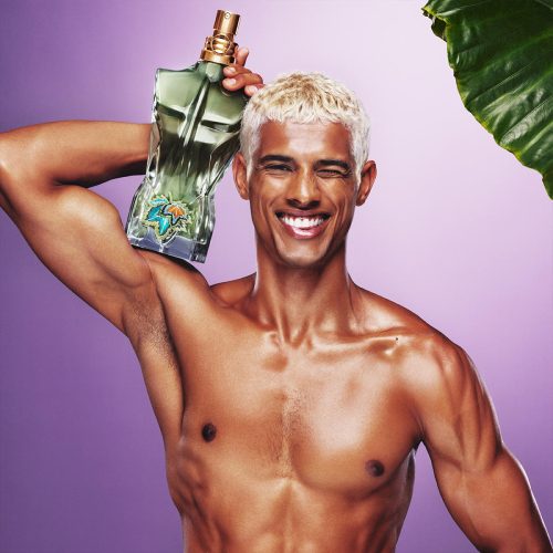 Jean Paul Gaultier Le Beau Paradise Garden muški parfem – egzotičan, svež i moderan miris prikazan u tropskom ambijentu koji simbolizuje luksuz, opuštenost i prirodnu muževnost. Zgodan momak, torzo muskarca na roze pozadini drzi parfem na ramenu